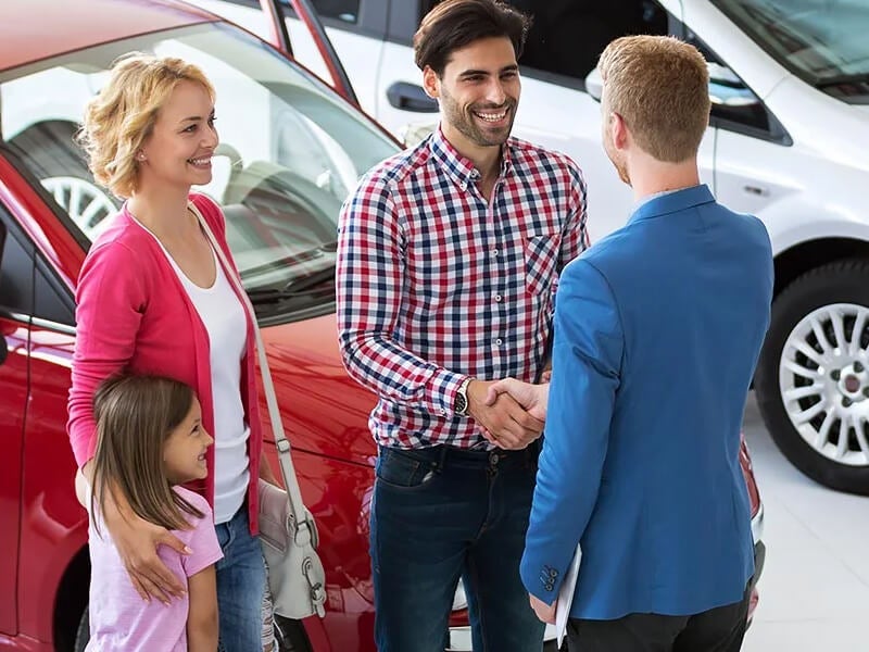 A salesman in a blue jacket shakes hands with a man, while a woman and child stand beside a red car.