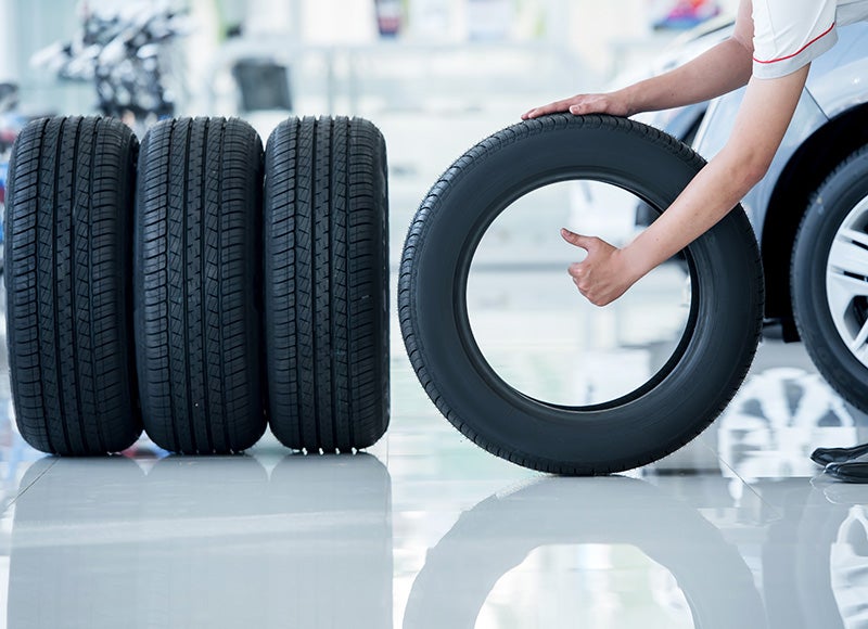 A person in a mechanic's uniform pushes a single new tire, giving a thumbs-up. Three other new tires stand beside it.