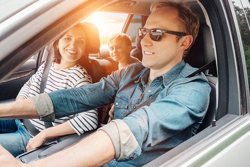 A family of three (father, mother, and child) sitting in a car, smiling. Sun shines through the window.