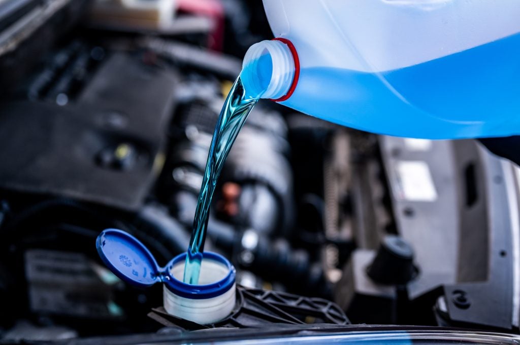 Blue windshield washer fluid pours from a bottle into a car's reservoir, visible over an open engine compartment.