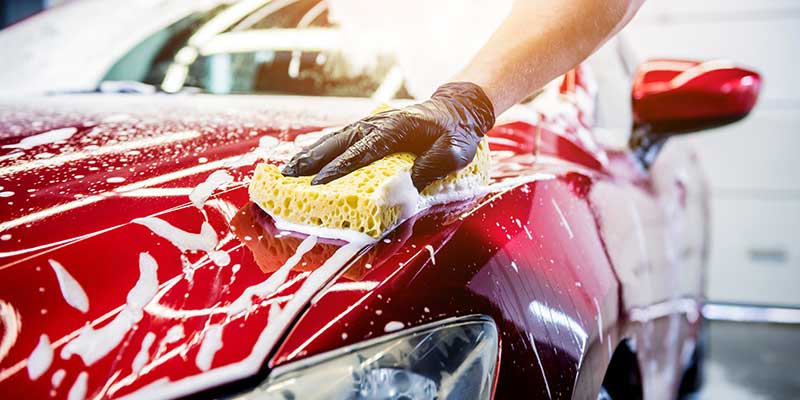 A person wearing black gloves washing a red car covered in white soap suds with a yellow sponge