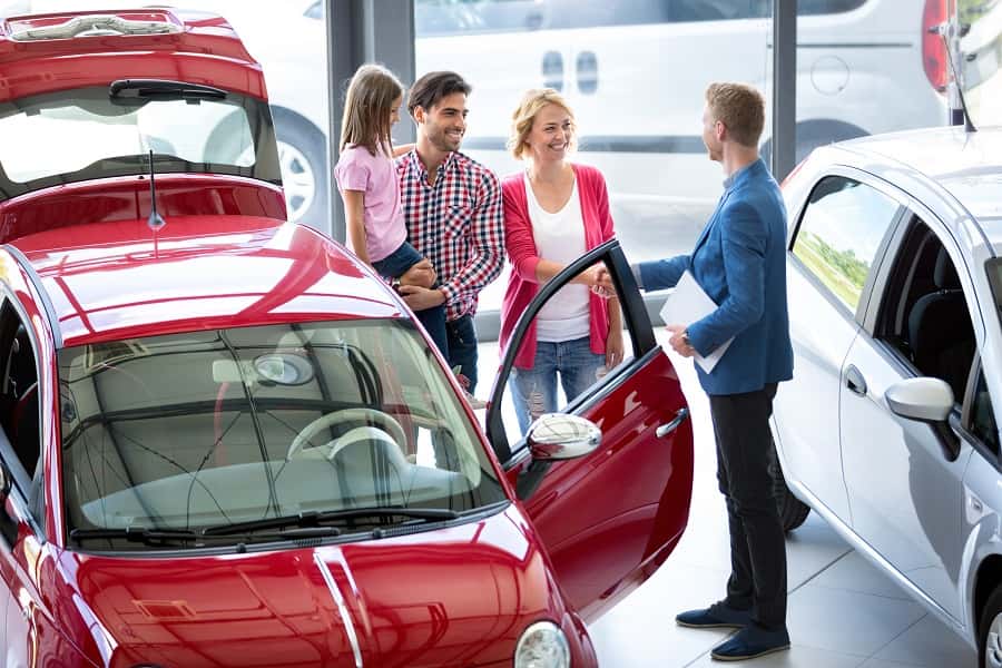 A family of three, an adult man, woman, and young girl, stands in a car showroom looking at a red car. A salesman talks to them.