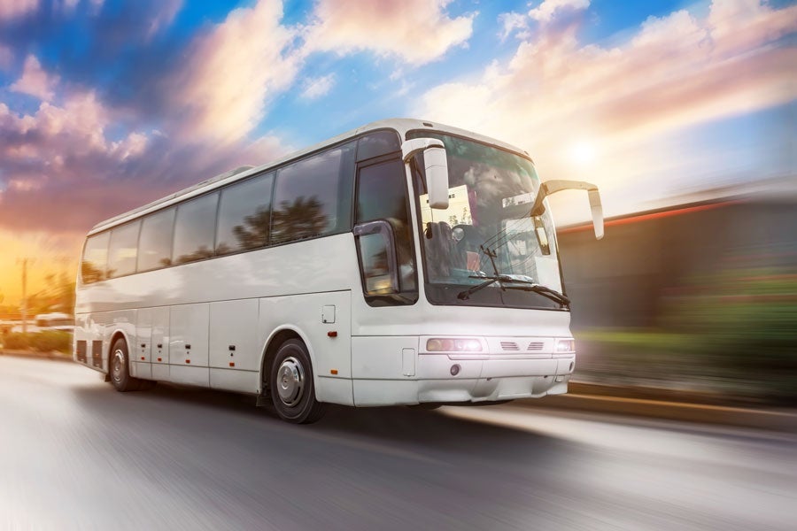 A white tour bus drives past with motion blur, creating a sense of speed. Blue and pink clouds are in the sky.