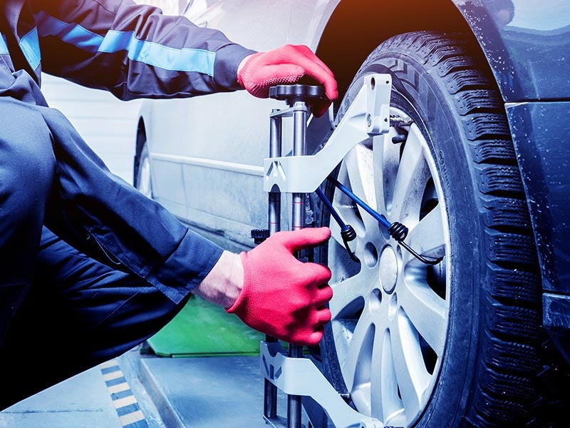 A mechanic in blue overalls and red gloves performs wheel alignment on a black car.