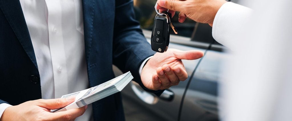 A person in a suit hands money to another person in exchange for car keys, symbolizing a transaction.