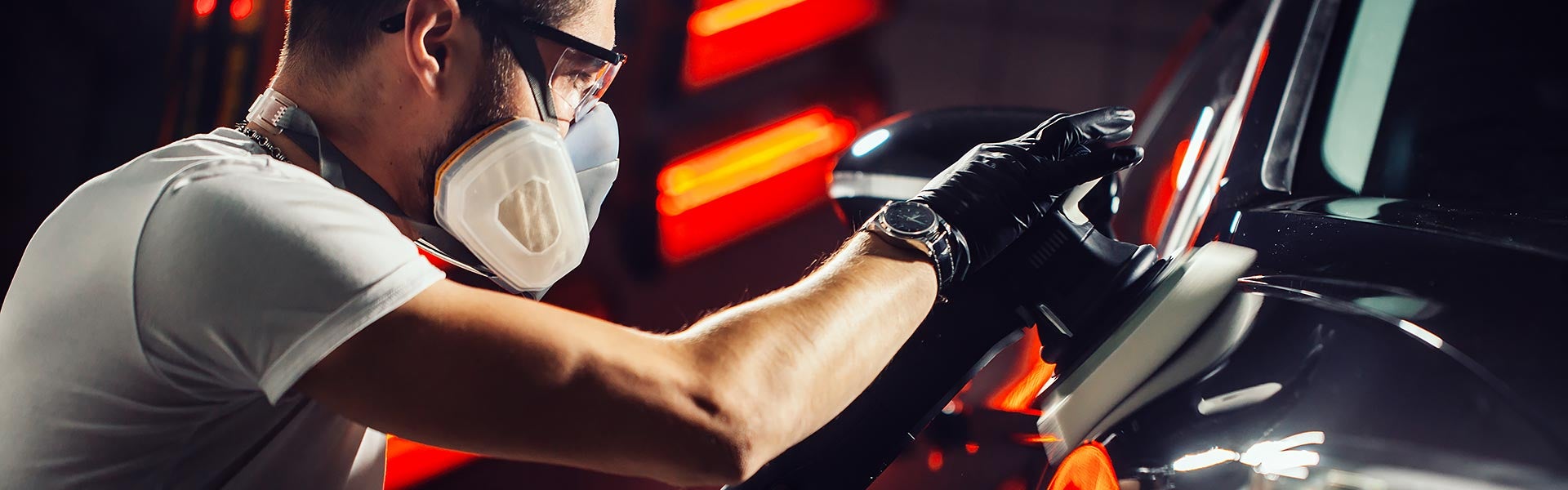 Person in a respirator mask and safety glasses polishing a dark car with a buffer.