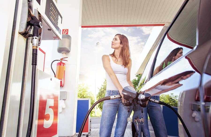 A young woman in a white tank top and blue jeans smiles while pumping gas into a dark car.