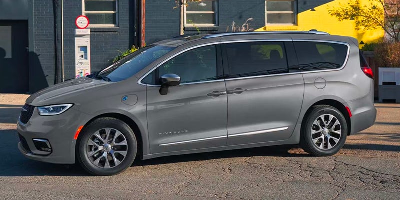 A gray Chrysler Pacifica Hybrid minivan parked on a cracked asphalt street, with a dark brick building in the background.