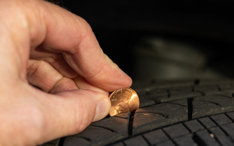A hand places a penny head-first into a tire's tread, demonstrating the penny test for tire wear.