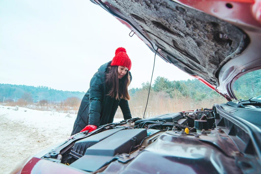 A woman in a red hat and black coat peers under the open hood of her broken-down car in a snowy winter landscape.