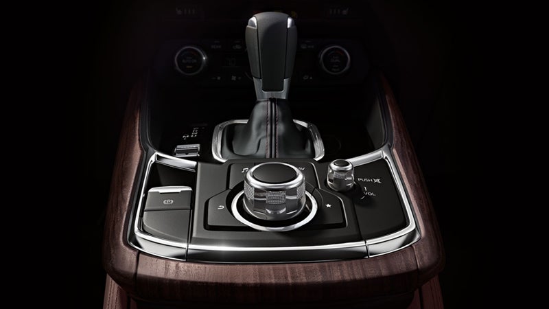 Console of a luxury car with a black leather gear shifter, a circular control knob, and brown wood trim.