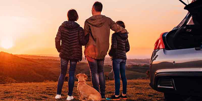 A family and their dog watch the sunset over a hilly landscape, with their car parked nearby.