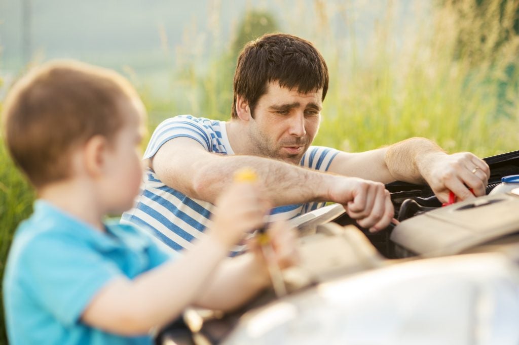 A man in a striped shirt leans over a car engine, hands working inside. A small child in a blue shirt stands in the blurry foreground.