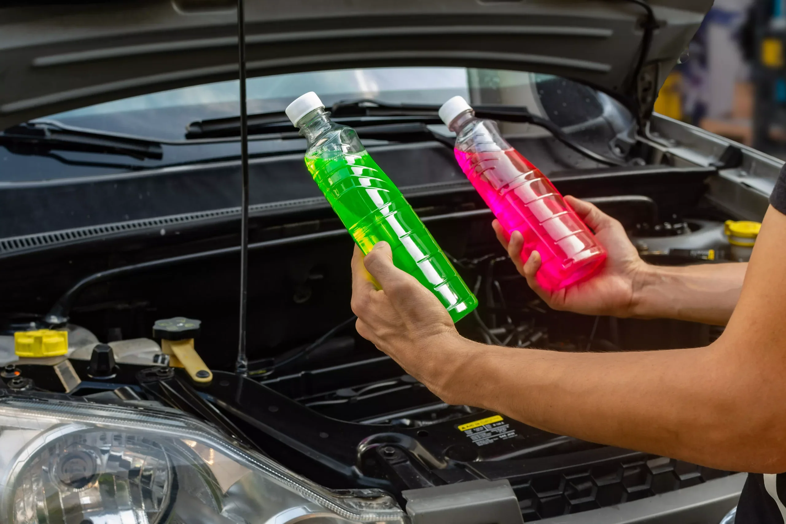 Hands holding bottles of green and pink car coolant over an open car hood.
