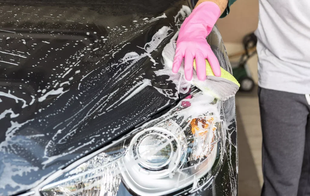 A person in a pink glove scrubs the headlight of a black car with a yellow and gray sponge, covered in soap suds.