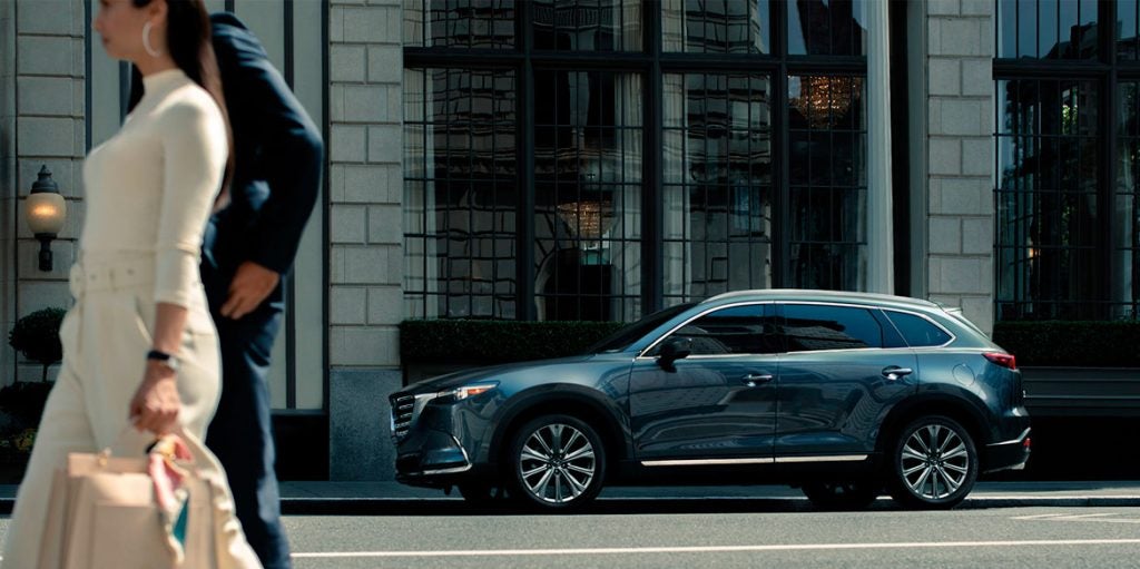 A dark gray Mazda CX-9 SUV is parked on a city street in front of a building with large windows.