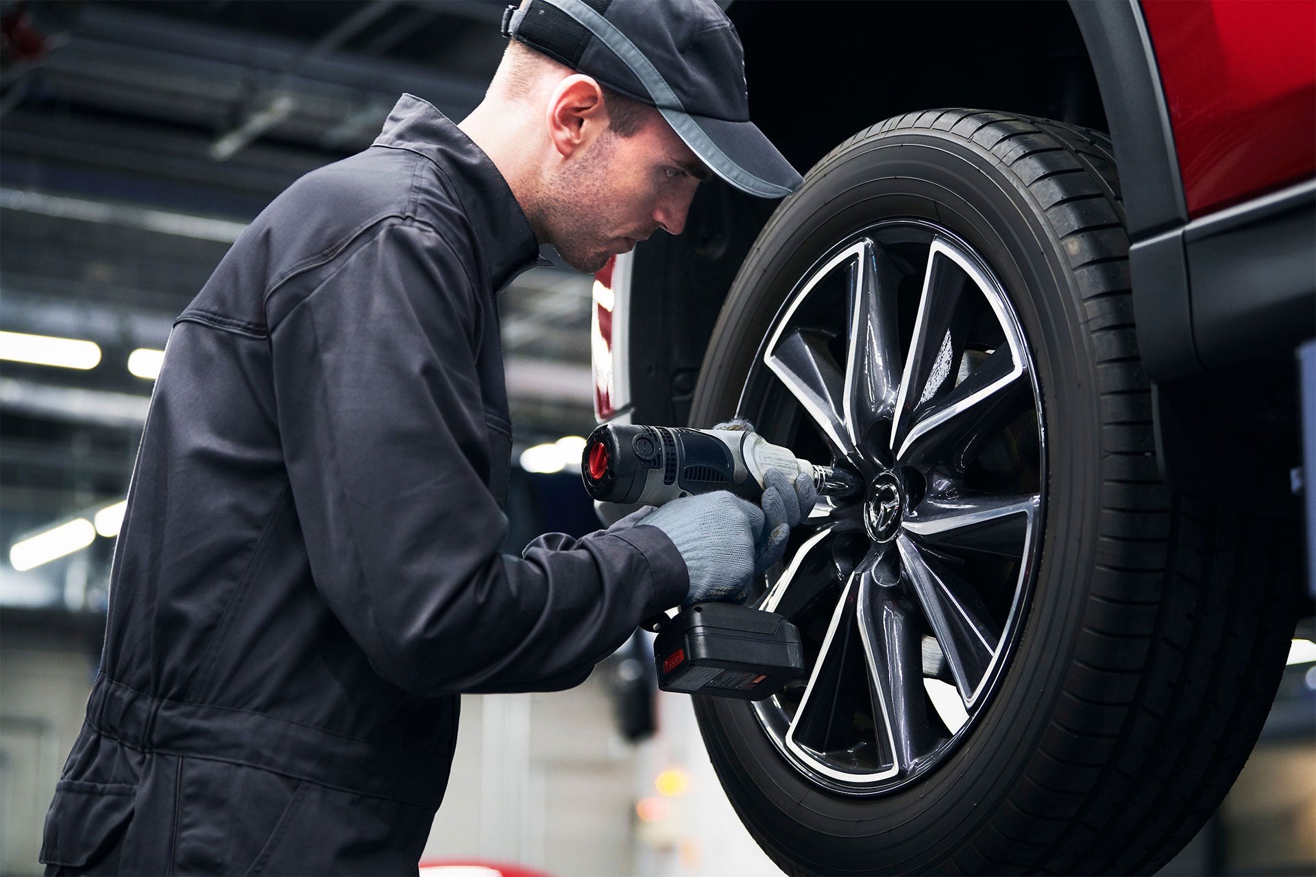 A mechanic in a cap and gloves uses an impact wrench to work on a car tire.