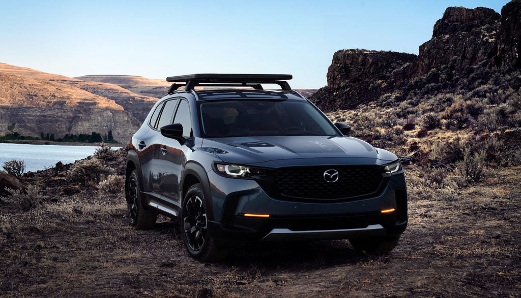 A dark gray Mazda CX-50 SUV with a roof rack sits on a dirt road backed by a lake and desert hills.