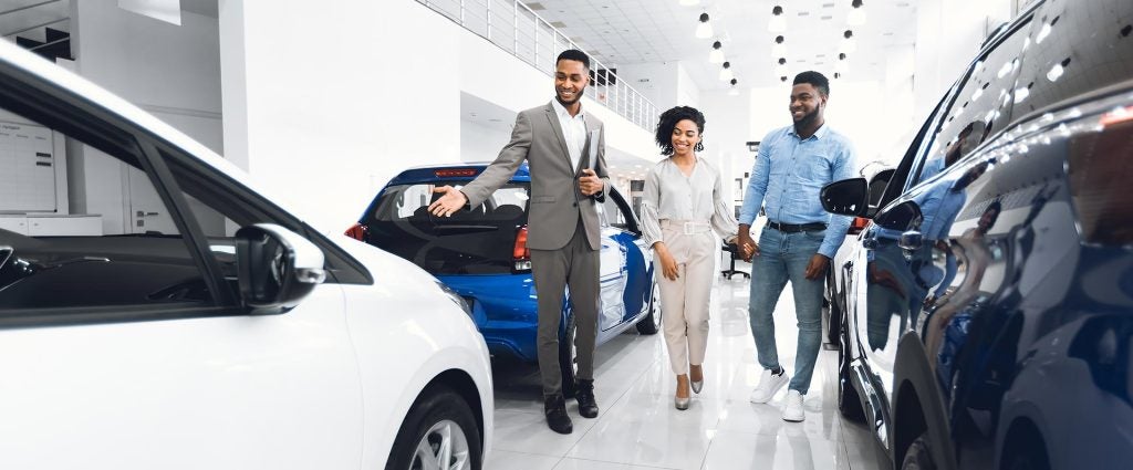A car salesman in a gray suit points at a blue car, smiling at a young couple holding hands in a car dealership.