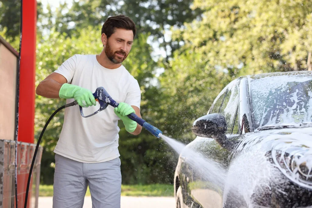 A man in a white tee shirt and green gloves uses a pressure washer to rinse soap off a black car.