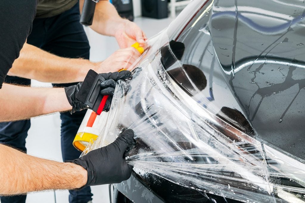 Two people apply clear protective film to a black car's headlight and hood, smoothing it with tools.