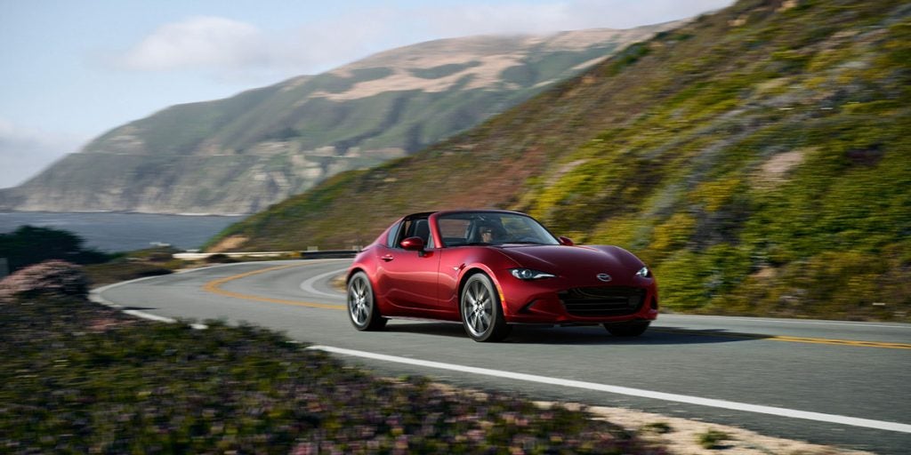 A red Mazda MX-5 Miata RF convertible driving on a winding coastal road with mountains and the ocean in the background.