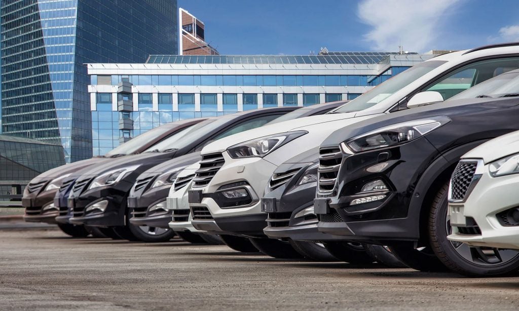 A row of parked cars, viewed close-up from the front corner, with tall modern buildings in the background.