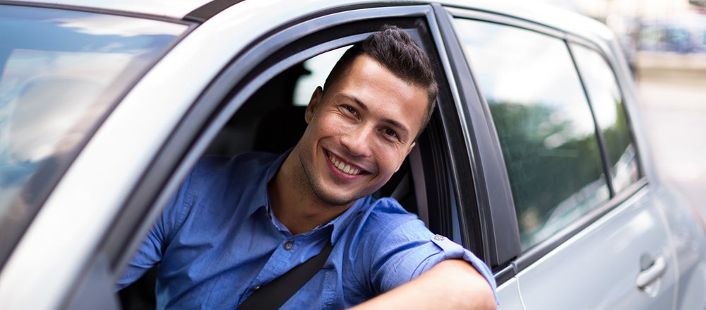 A smiling man in a blue shirt with his seatbelt on leans out the window of a light-colored car.