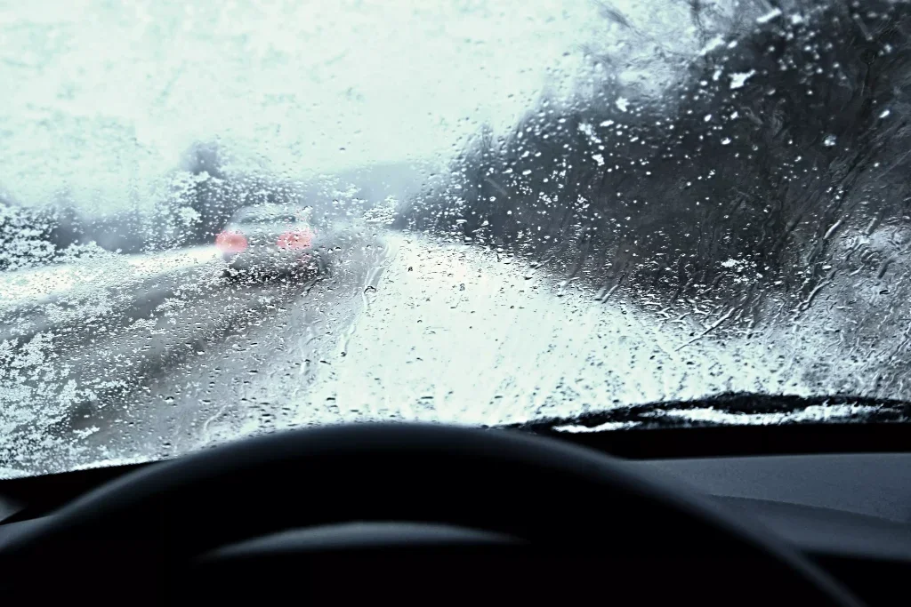 View from inside a car on a snowy road, looking through a snow-covered windshield at a grey sky.