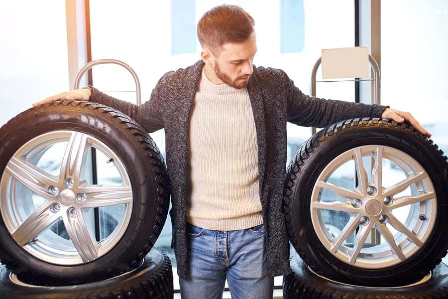 A mechanic in blue work clothes and gray gloves rolls a tire with a black and silver rim across a gray floor.