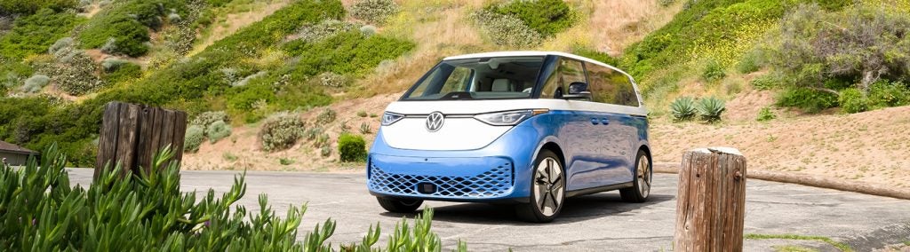 A blue and white volkswagen parked on asphalt in front of a desert looking background surrounded by greenery 