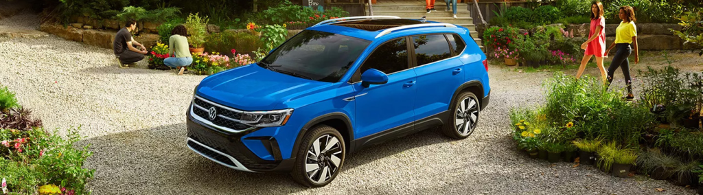 A blue volkswagen taos sits in a garden type area and two women are walking by looking at it 