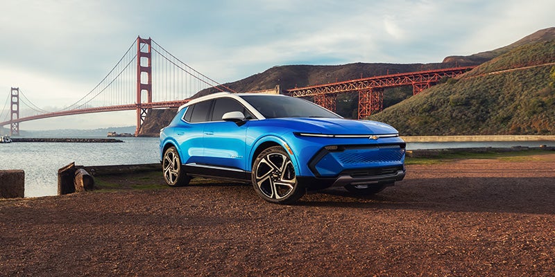 Blue Chevrolet Equinox EV parked on a gravel, showcasing a modern electric crossover design with scenic coastal and mountain views in the background.