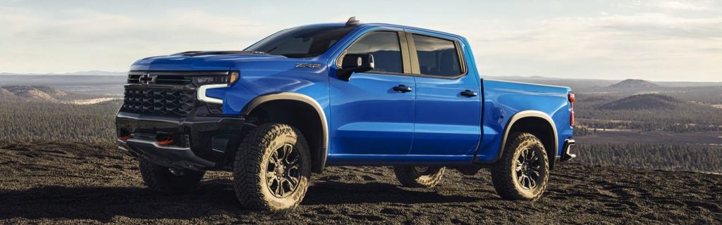A blue Chevrolet Silverado 1500 pickup truck parked on rocky terrain with a mountainous background.