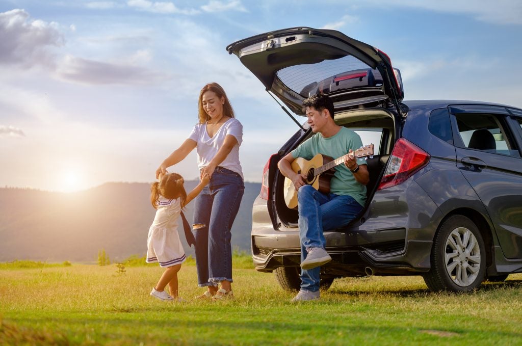Family enjoying outdoor moment by an SUV with open trunk, with a man playing guitar while a woman and child interact in a sunny field at sunset.