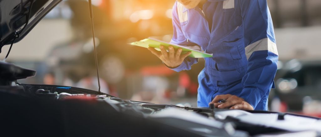 Automotive technician inspecting a vehicle engine bay while holding a tablet, performing diagnostics in a service garage with warm sunlight in the background.