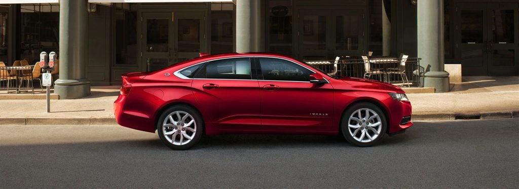 A red Chevrolet car parked on the side of a street, in front of a cafe with outdoor seating.