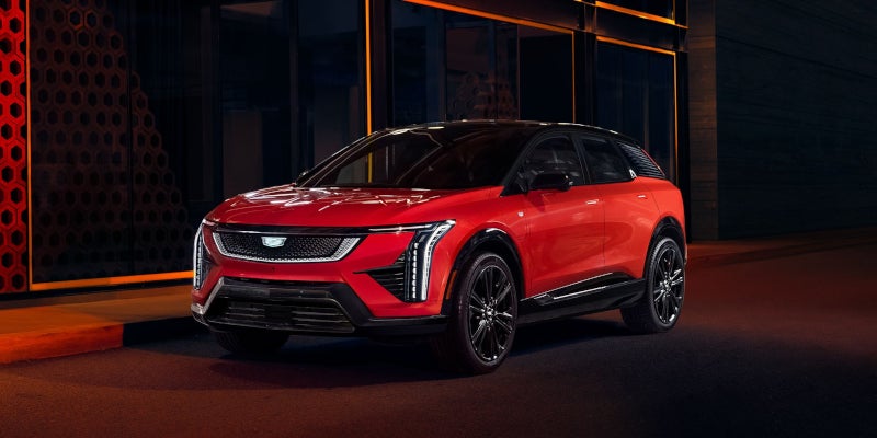 Front-three-quarter view of a red Cadillac OPTIQ with a black roof, parked at night in front of a modern building with glowing orange accents.