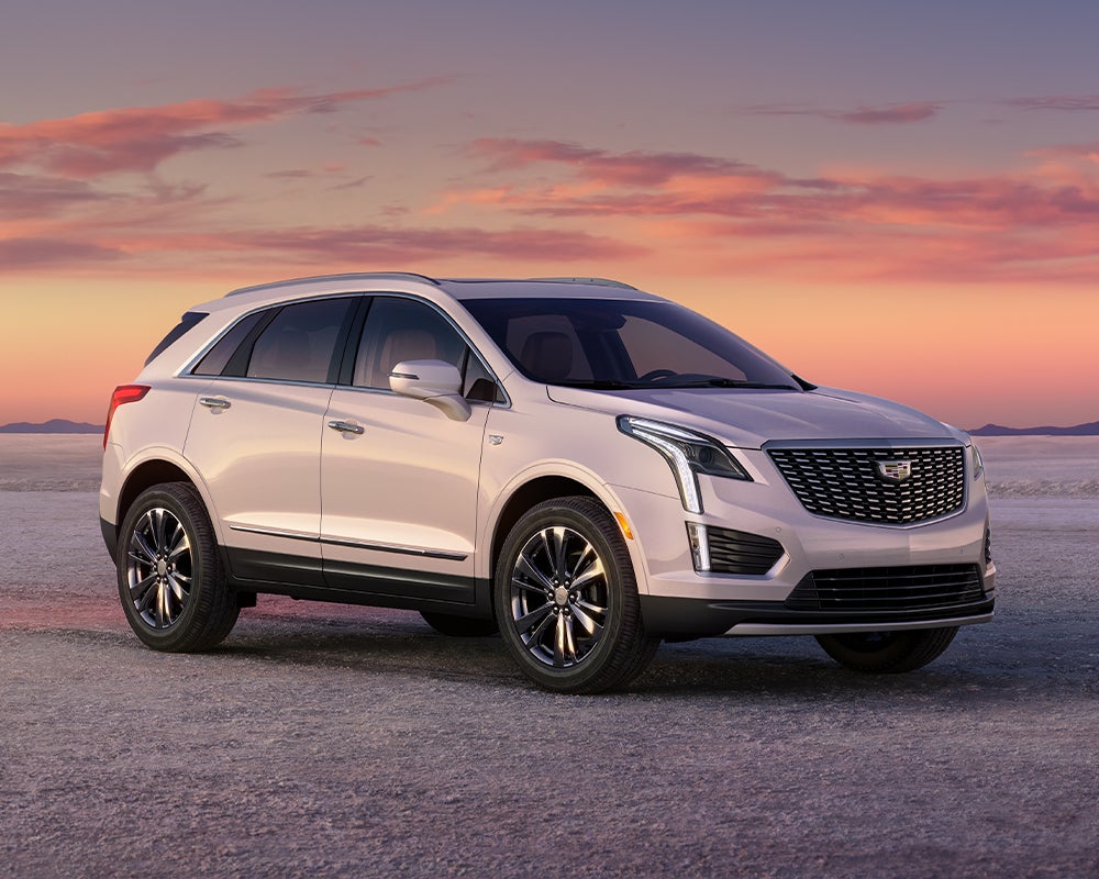A white Cadillac XT5 parked on a flat, open desert landscape under a vibrant pink and orange sunset sky.