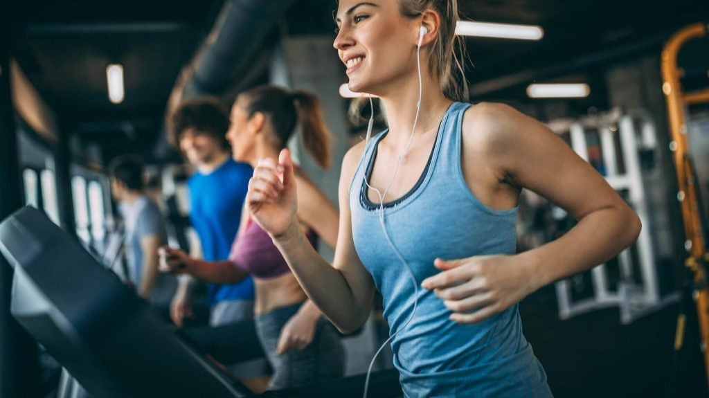 a blonde woman jogs on the treadmill with earphones in, next to a few other people who are also jogging on the treadmill 