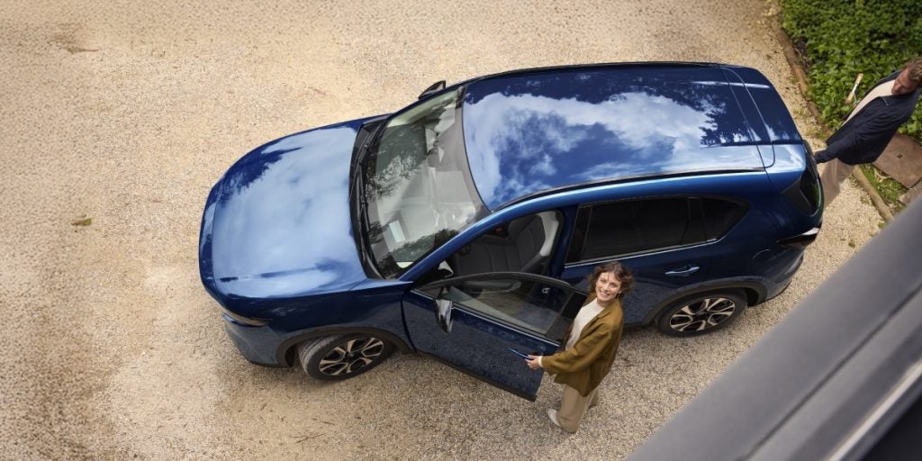 Top-down view of a blue SUV parked in a driveway with the driver’s door open, showing a person standing beside the vehicle looking up at the camera.