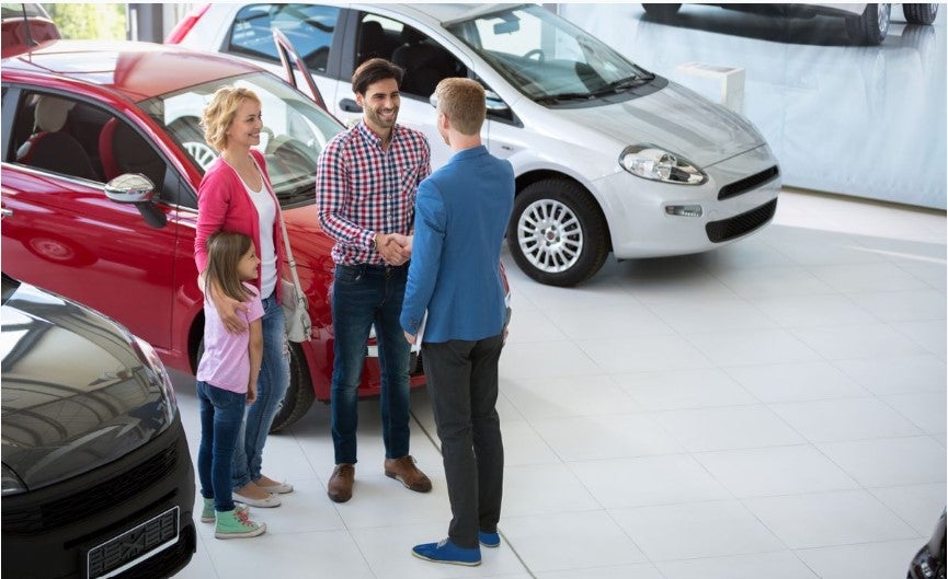 Family of three shaking hands with a car salesperson inside a car dealership showroom, surrounded by parked vehicles, suggesting a vehicle purchase or agreement discussion.