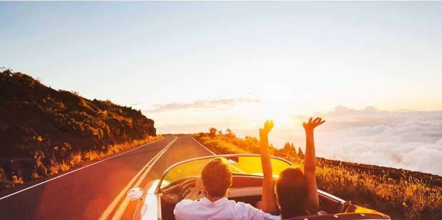 Couple in a convertible car driving along a scenic coastal mountain road at sunset, with warm sunlight in the background as they raise their arms in celebration.