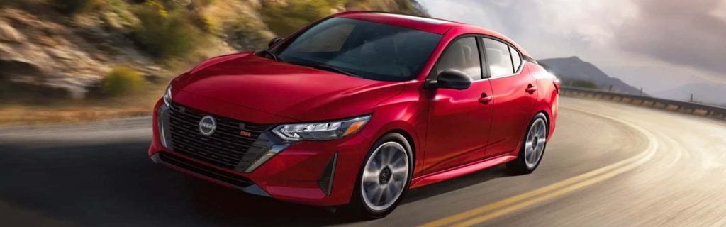 Red 2026 Nissan Sentra sedan driving on a winding mountain road, showcasing its sporty styling and efficient compact performance