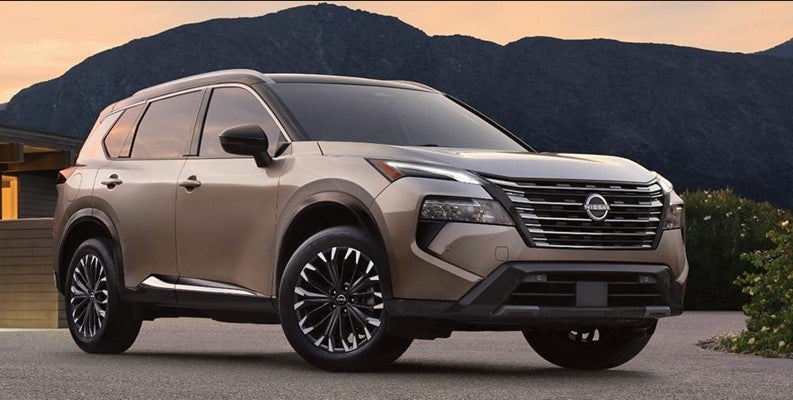 Front three-quarter view of a tan Nissan Rogue parked outdoors at sunset, with mountains in the background and a modern residential setting nearby.