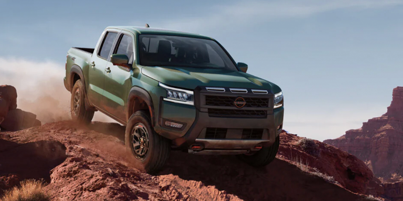 Green pickup truck driving down a rocky off-road trail in a desert canyon, kicking up dust while navigating a steep incline.