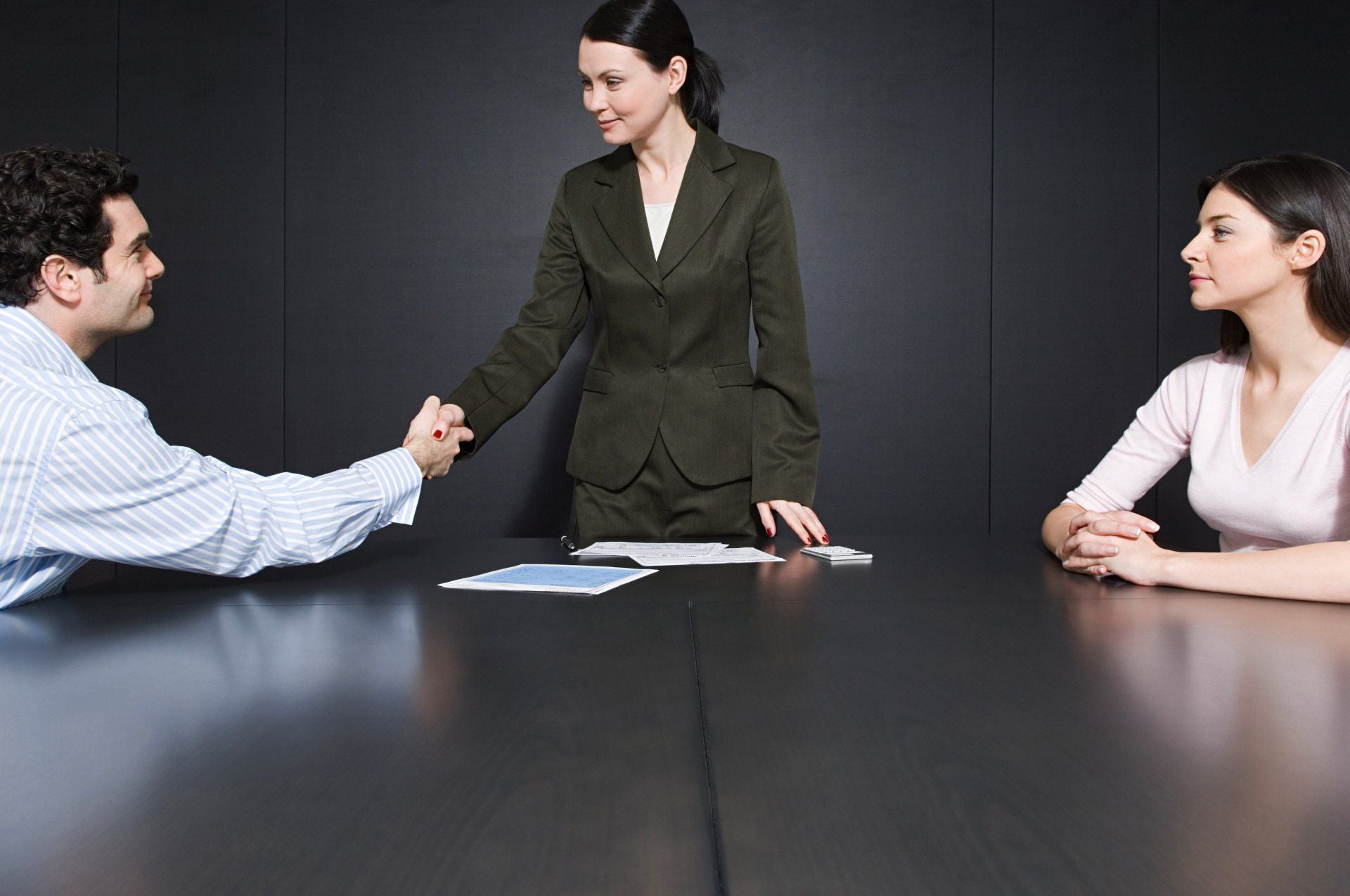 a woman in a black suit shakes hands with a man in a blue shirt sitting opposite a woman in a pink shirt