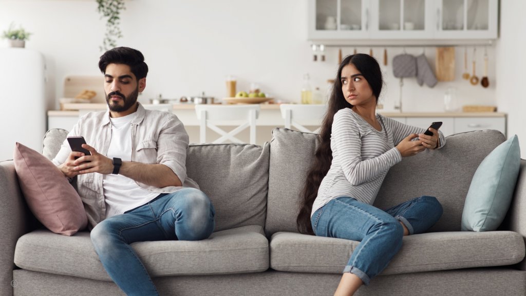 A couple sits on a couch with their backs to eachother, while texting