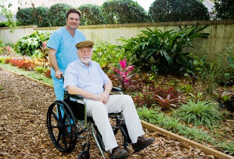 A nurse pushes an elderly man in a wheelchair through a garden.