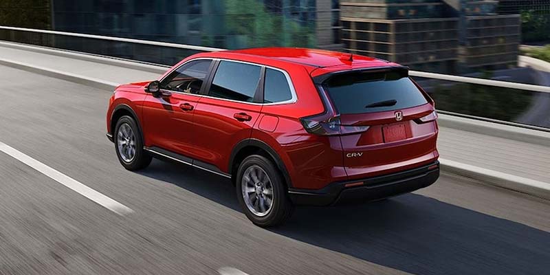 A red 2026 Honda CR-V Hybrid gliding through a city road, with tall buildings lining the horizon
